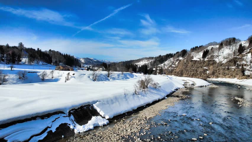 Drone aerial footage of the winter landscape surrounding the Shinano River in Niigata Prefecture, Japan. The video captures snow-covered rural villages, winding rivers, mountain valleys, forests, and peaceful countryside scenery from above. The contrast between flowing water and deep snow creates a cinematic atmosphere unique to Japan’s snowy regions. Suitable for travel, tourism, rural landscapes, environment, geography, winter scenery, and nature-themed projects.