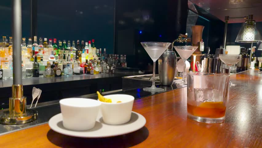 A whiskey cocktail sits on a wooden bar counter alongside snacks in a luxury lounge