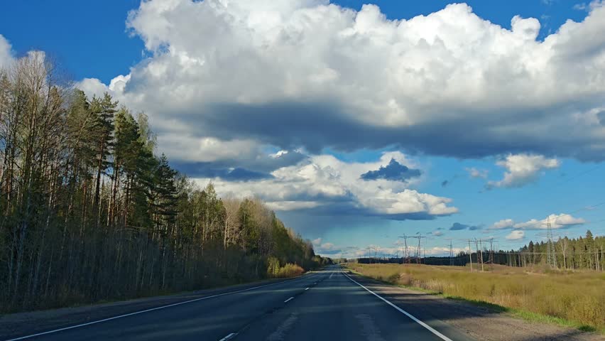 A country road leads towards the horizon flanked by fields and trees under a dramatic sky with large white clouds. rural travel, open landscape, road trip.