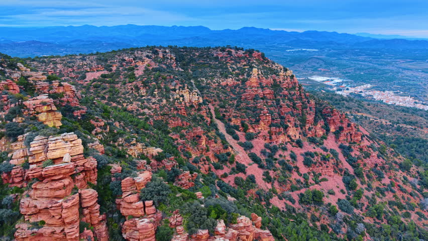 Panoramic aerial view of red rocky mountains and distant horizon. Wide landscape showing red sandstone slopes with green shrubs and blue mountains in the distance.