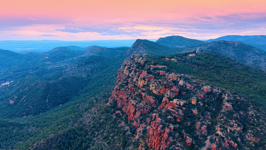Vibrant sunset sky over red sandstone canyon and mountain forest. Wide aerial shot of red rock cliffs at dusk with a pink and orange sunset sky over a forest valley and mountains.