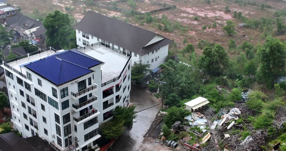 Aerial view of a very windy rainstorm over modern white buildings with a blue roof, featuring swaying trees and cars on a wet street.