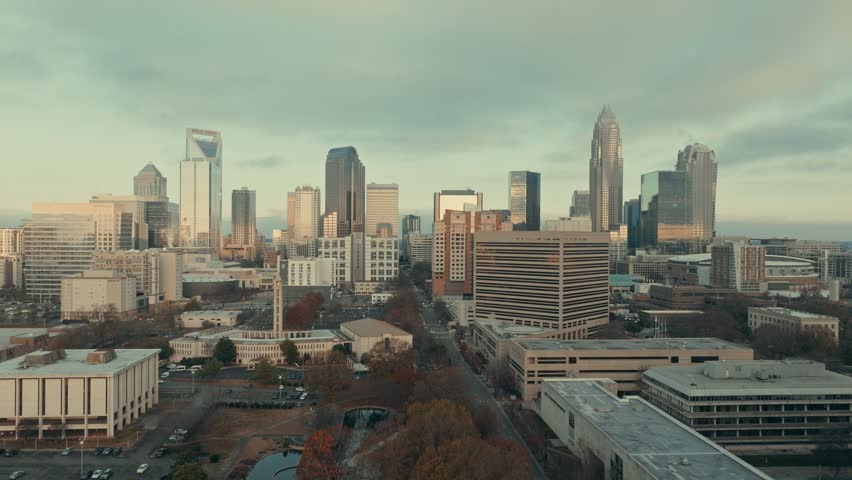 Cinematic aerial rise revealing panoramic Charlotte North Carolina skyline with glass skyscrapers catching warm golden light through moody overcast sky and autumn foliage below