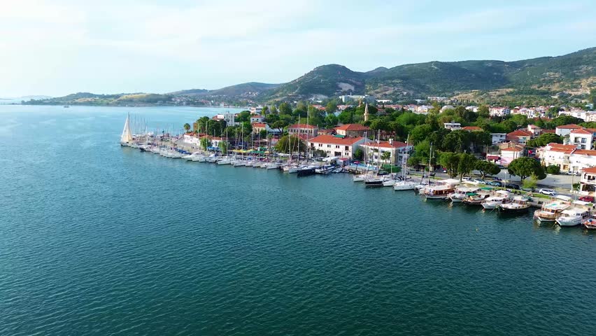 An aerial perspective captures a vibrant coastal town with a harbor packed with sailboats and yachts, set against a backdrop of lush green hills. Turkey, Izmir, Eski Foca