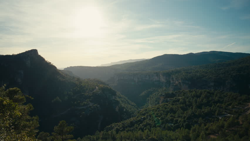 Late sunlight spills across a broad canyon basin in Catalonia, Spain, where dark cliff walls, wooded slopes, and distant plateaus create a quiet and dramatic sense of depth.