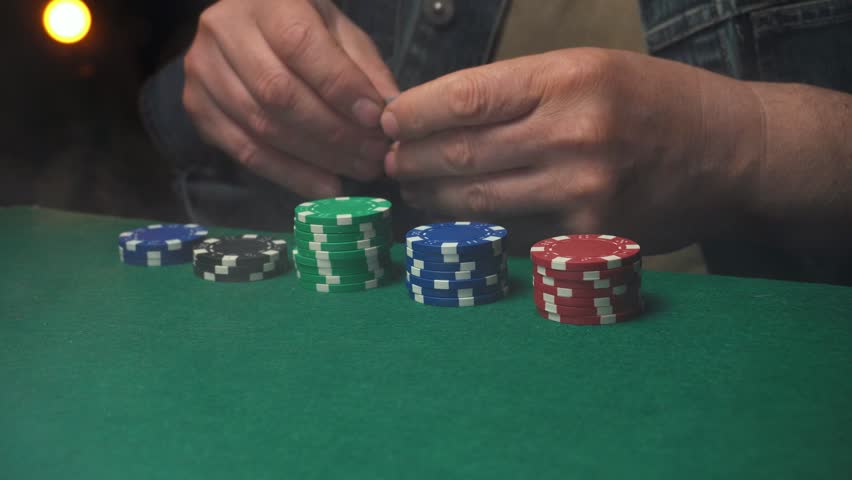 Male poker player in denim jacket lays down playing cards on a green felt table, with various colored poker chips stacked nearby in a cozy, low-light gaming atmosphere