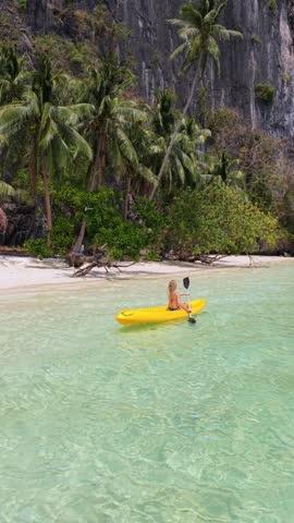 Woman kayaking in crystal clear turquoise water near tropical beach in El Nido, Palawan, Philippines. Exotic island scenery with limestone cliffs, palm trees, and paradise coastline.