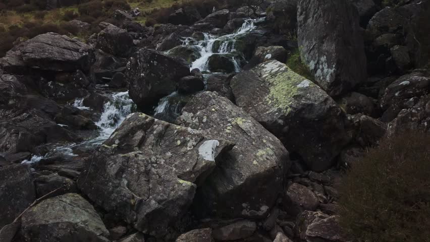 A scenic opening shot of water crashing over stones, transitioning into a view of the rugged hiking path leading toward Llyn Idwal under a misty mountain sky.