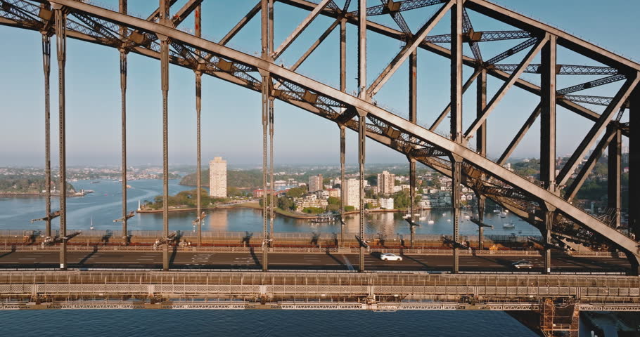 Sydney Harbour Bridge structure framing a view of urban cityscape with cars traveling on road, with water and buildings in the background, representing travel and exploration in Australia. Aerial shot