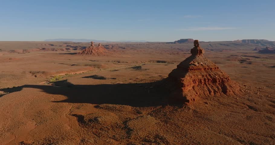 Drone tracks sideways past Rooster Butte at Valley of the Gods, Utah, revealing a vast red desert landscape with distant formations and mountains under clear evening skies.