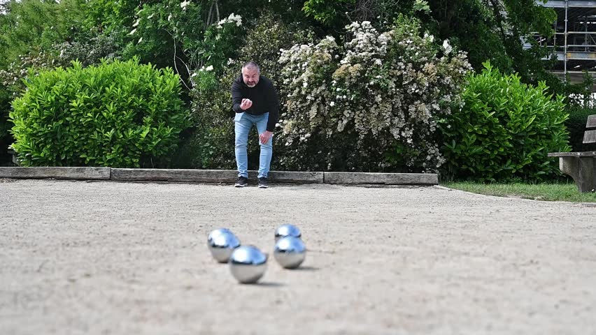 Man playing petanque on sunny park court with boules scattered on gravel, casual outdoor leisure and recreation game in European setting
