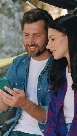 Joyful young adult couple man and woman sitting in car trunk with children on background using tapping on phone while laughing. Mother and father surfing internet on cellphone. Family road trip with