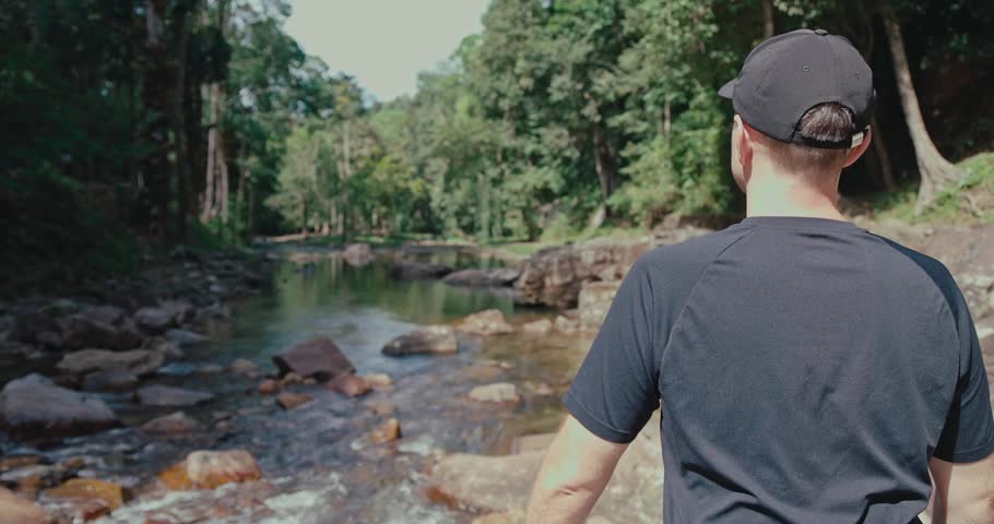 Man in black t-shirt and cap stands by rocky river in lush tropical forest. Back view of male traveler exploring nature during sunny day. Adventure and outdoor lifestyle.