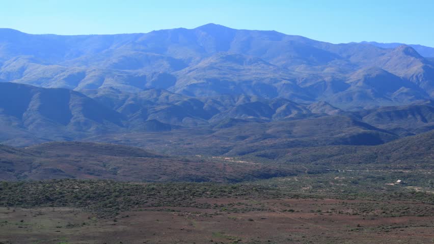 Valley in the foothills of Arizona, high-elevation desert zone within the Sonoran Desert. The image shows a deep valley and stratified mountain ranges - a landscape characteristic of basin-and-range topography or the protected Sierra Ancha Wilderness Area. In the valley, a small cluster of buildings from a remote settlement is visible. The slopes of the hills are covered with sparse, drought-adapted shrub vegetation, including chaparral and desert shrubs.