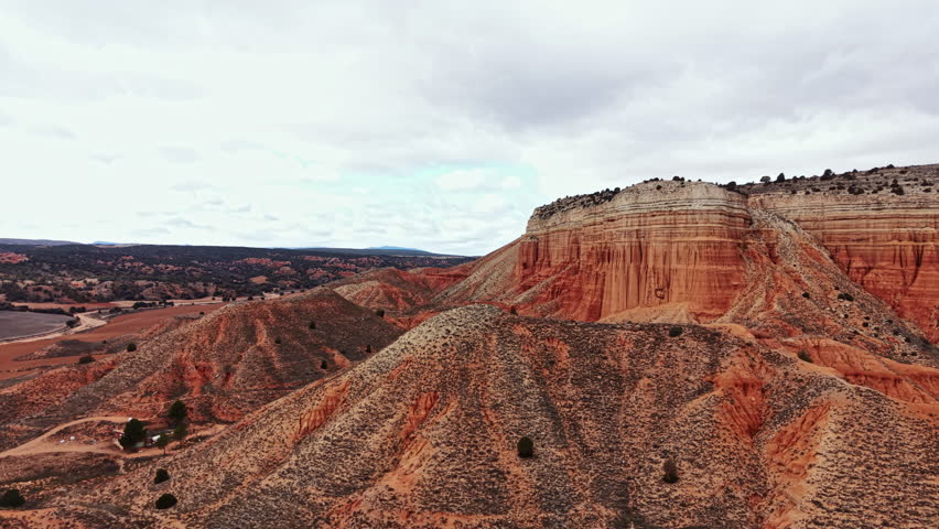 A large canyon stretches across the land under a cloudy sky. The terrain shows layers of rock and dry plants. The view highlights the rugged landscape and distant hills.