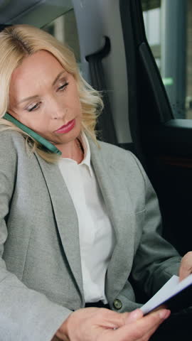 Beautiful business woman in glasses sitting on backseat of expensive car when working, talking on phone while checking important documents holding in hands. Successful career development, freelancer