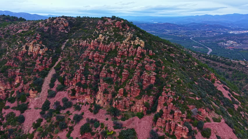 Rocky hills rise from the ground with sparse trees scattered around. A valley is visible in the distance, along with other mountain ranges under a cloudy sky.