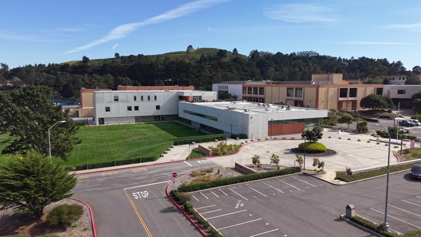 Sunny afternoon aerial view of Skyline College campus, including modern buildings, a large parking lot, and green hills in the background, located in the city of San Bruno, California