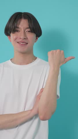 Young smiling man making a thumbs-up gesture to the side against a turquoise studio background with copy space.