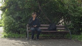young man checking phone on bench with backpack, adjusting earbuds and scanning messages, glancing toward gravel path and leafy hedge, soft daylight and calm commuter energy, subtle optimism - Powered by Shutterstock - Get 15% off with code: PIKWIZARD15
