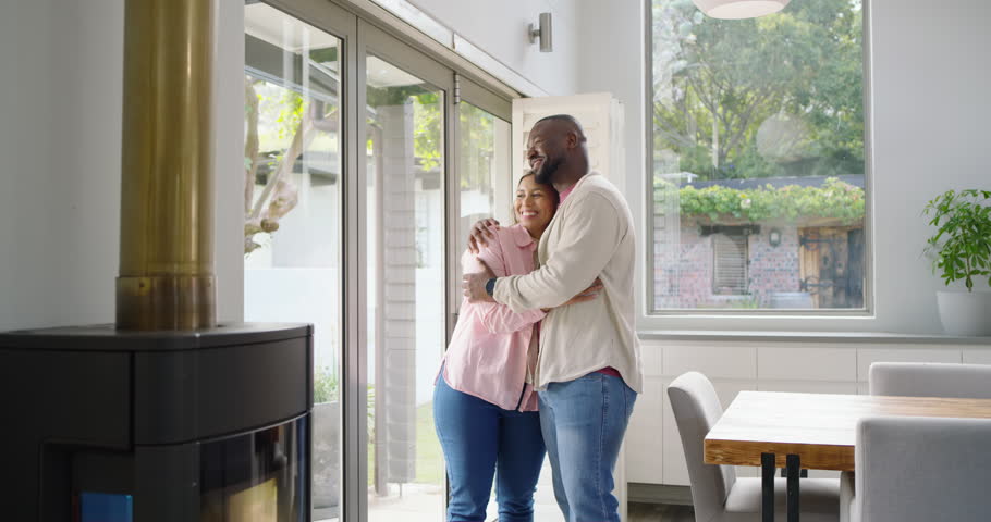 African American couple standing by glass doors, embracing, woman pointing outside to admire yard. Sunlit, interior, openplan, kitchen, dining, fireplace, plant