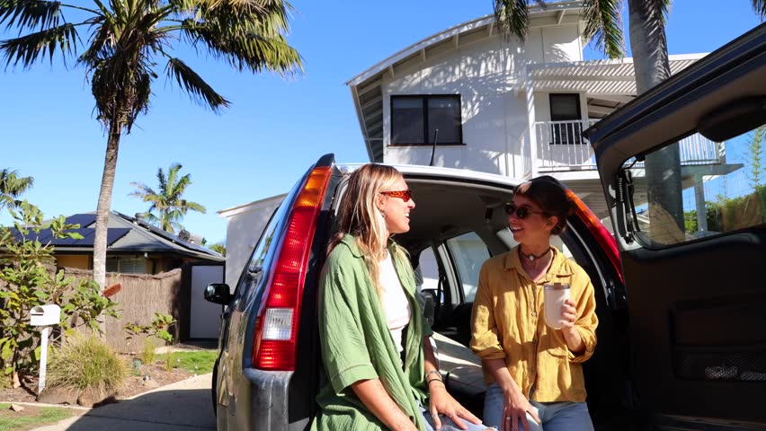 Two cheerful young women sitting in the open trunk of a car, laughing and chatting before going surfing on a sunny day in australia, embodying a carefree and adventurous lifestyle