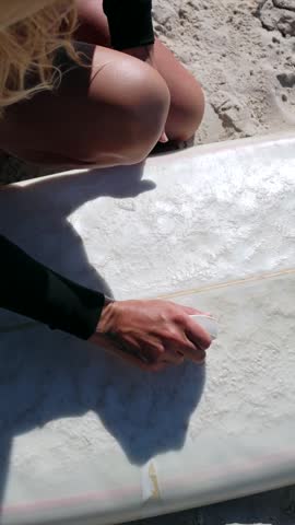 Blonde woman in a wetsuit preparing for surfing by applying wax to her surfboard on a sunny day at the beach, getting ready for a water sports session in australia