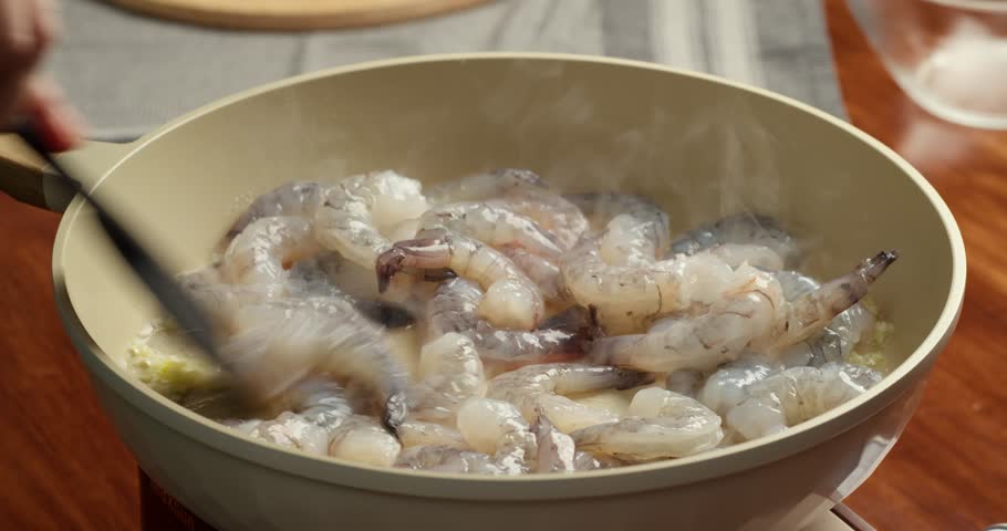 Close up of raw shrimp being cooked with garlic and oil in a frying pan during home cooking preparation. Fresh seafood frying in hot pan with steam in kitchen.
