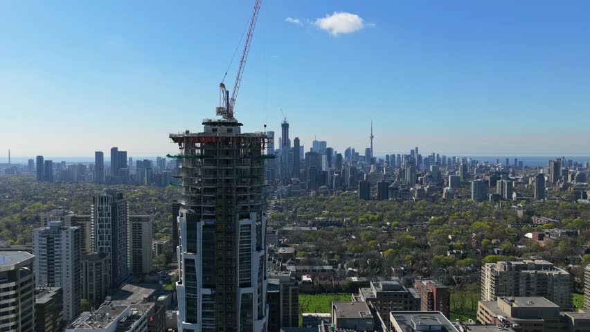 High rise skyscraper construction towering over midtown Toronto during day, downtown core skyline, CN Tower, Lake Ontario on horizon under clear blue sky
