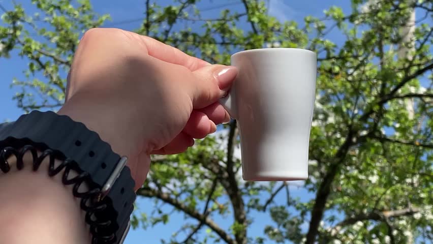 Hand holding white ceramic cup of coffee against green sunny trees.