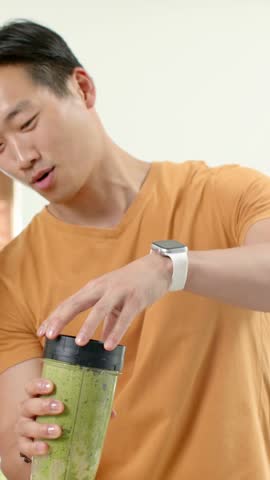Vertical video: Blender stopping Asian man in orange shirt twisting lid off in kitchen, showing cup. Demonstration, appliance, smoothie, home, countertop, modern, vertical video