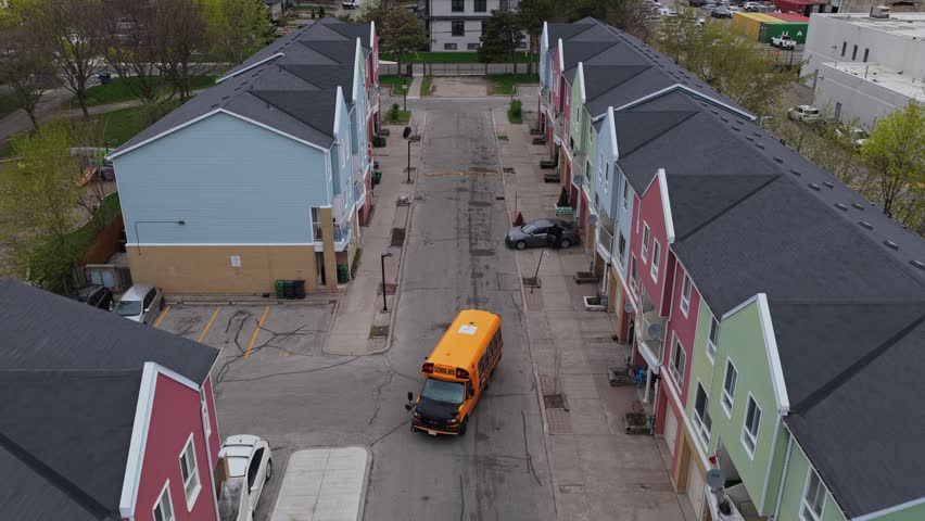 Yellow school bus reversing on residential street between modern townhouses in Canada revealing city skyline with aerial perspective backing away and rising