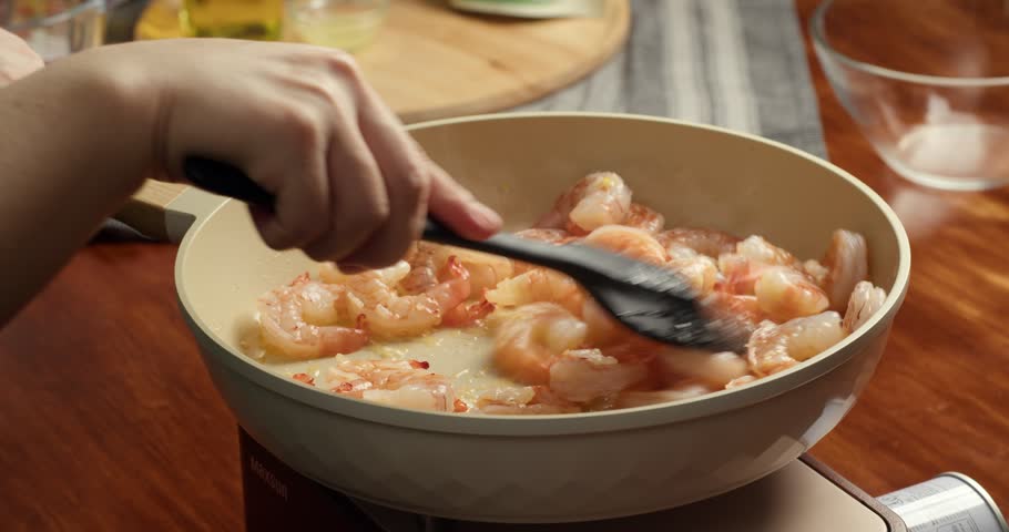 Close up of chef stirring cooked orange shrimp with silicone spatula in a frying pan during home cooking preparation in kitchen. Hot seafood dish being prepared on stovetop.