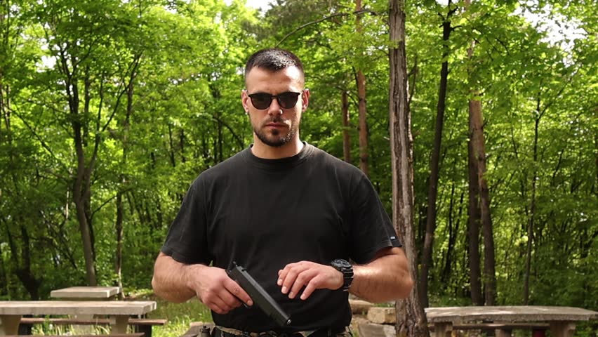 Focused man in sunglasses raising and aiming a black airsoft pistol in a forest clearing, preparing for target shooting practice outdoors with tactical stance and natural green background.