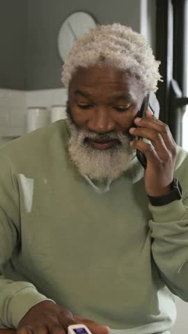 Vertical video: Answering call, senior man holding phone, checking white box at kitchen counter. Device, gadget, smartwatch, technology, home, interior, vertical video