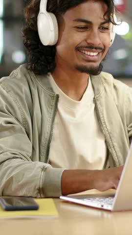 Vertical video: Typing man in khaki jacket, white headset, starting work in cafe with laptop, phone. Headphones, notebook, desk, smartphone, coworking, workspace, vertical video