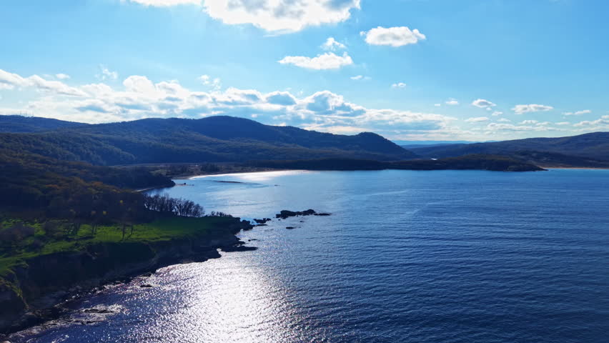 A stunning coastal scene features gentle waves lapping against rugged rocks, surrounded by lush green hills while fluffy clouds drift across the bright blue sky.
