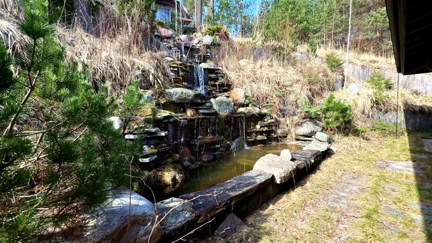 Small man made waterfall with tiered stone steps cascading into a backyard pond in early spring