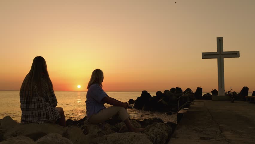 Two women watching sunrise, silhouette by ocean, cross on pier.