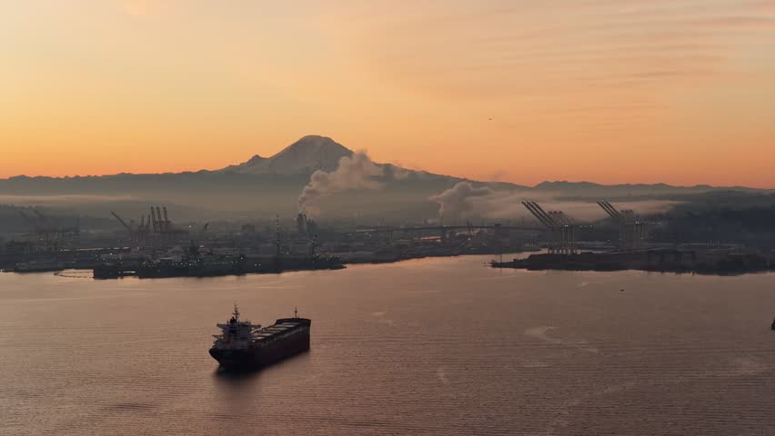 Mount Rainier above Seattle docks at sunrise with cargo ship, port cranes and calm harbor water under orange sky. g.