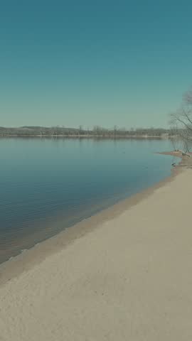 Aerial vertical rise from sandy St Croix River beach in downtown Hudson Wisconsin revealing calm glassy water, tree-lined shores, mid-river island and rolling hills beyond