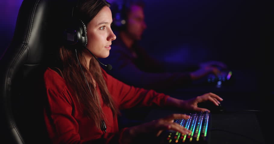 Closeup of Caucasian young adult female gamer in headset sits at computer, playing online video game in cybercafe. Competitive esports, professional gaming, leisure entertainment technology concept