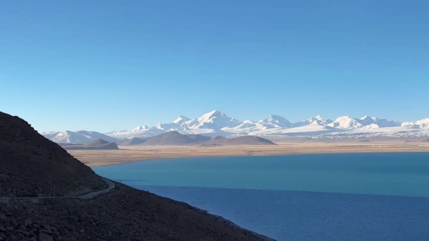 Wide panoramic view of a winding road along the turquoise Puma Yumco lake with majestic Himalayas snow peaks in Tibet.