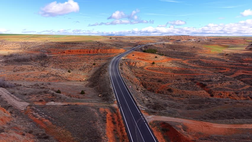 Two-lane road winding through red clay badlands and arid scrubland under a blue sky with scattered clouds, stretching toward a distant green horizon