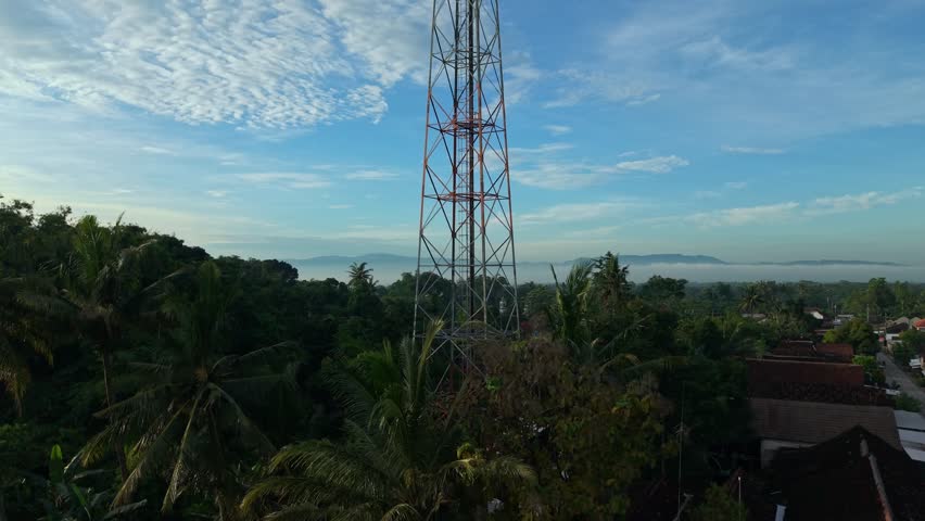 Aerial view of a tall telecommunications tower surrounded by lush green palm trees and rural houses under a blue sky in Kemalang, Central Java, Indonesia.