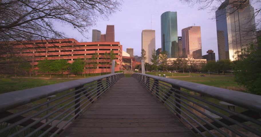 Smooth camera motion over a pedestrian bridge overlooking the houston downtown skyline with lush green parks and contemporary buildings under a clear sky, offering a dynamic urban and natural blend.