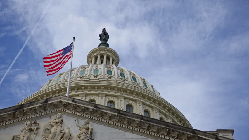 American flag waving proudly on Capitol. American flag under blue clouds over US Capitol. American flag patriotic symbol of Amerika. American flag on iconic Capitol dome in Washington DC.