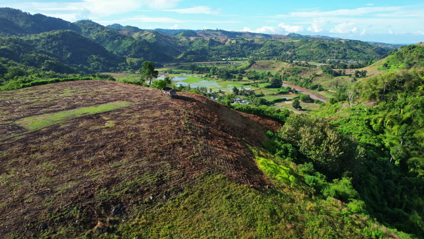 A hill covered with dry harvested corn crops in the Lunyuk area of Sumbawa Regency, Indonesia, seen in aerial drone footage with a small farmer hut on the hillside, while a lush green valley, a brown natural river, and Plara local village stretch across the background.
