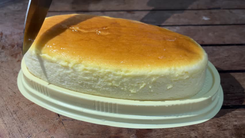 Close up footage of a sharp knife slicing through a soft fluffy Japanese cheesecake on a plastic base over a rustic wooden table background