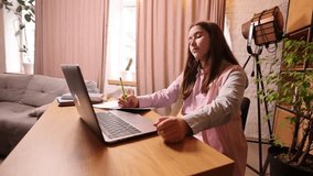 Teen girl sitting at desk at home in brightly lit living room, taking notes during her online lesson with teacher on laptop. Concept of remote learning, education - Powered by Shutterstock - Get 15% off with code: PIKWIZARD15